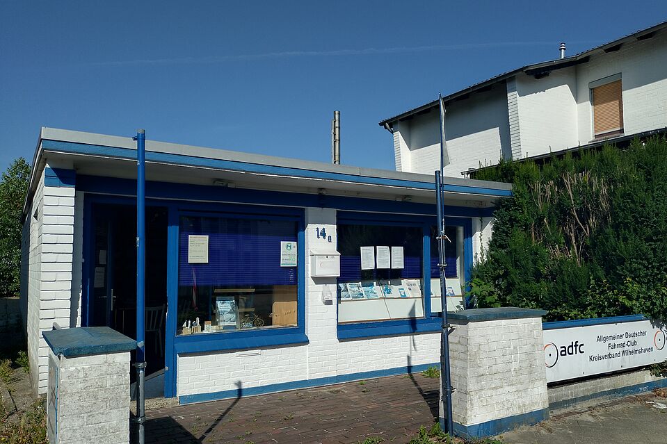 Infoladen Ein weißer, gemauerter Bungalow mit blauen Tür- und Fensterrahmen.